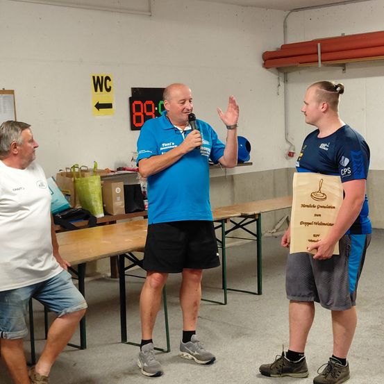 Drei Männer stehen in einem Raum, einer hält ein Mikrofon und spricht, ein anderer hält ein Zertifikat. Der Raum hat Tische, ein Schild mit 'WC' und eine Digitaluhr mit 88,6.