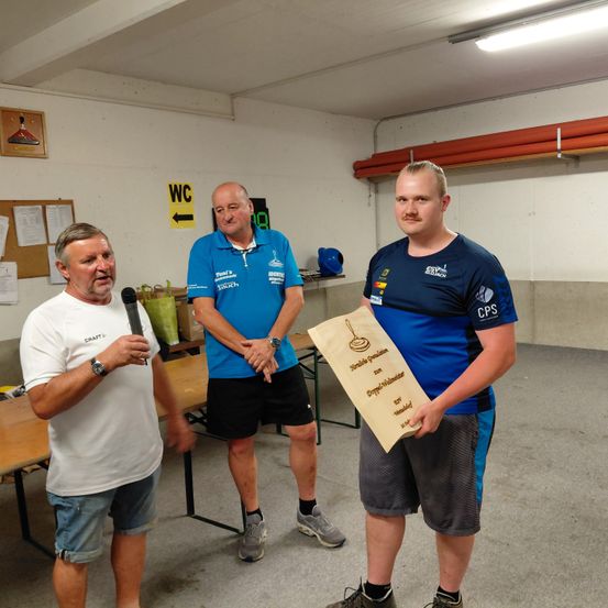 Drei Männer stehen in einem Raum, einer hält ein Mikrofon, ein anderer ein Zertifikat und der dritte trägt ein blaues T-Shirt. Hinter ihnen steht ein Tisch mit Papieren und ein Schild, das auf die Toilette zeigt.