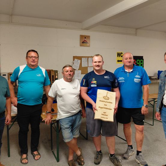 Fünf Männer in Sportbekleidung stehen in einem Raum. Ein Mann hält ein Zertifikat und sie scheinen alle für ein Foto zu posieren. Ein Tisch mit einer Flasche steht hinter ihnen.