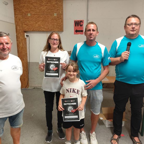 Fünf Erwachsene stehen in einem Raum, zwei Frauen und drei Männer, halten Trophäen. Die Frauen tragen eine Brille und Turnschuhe. Das Mädchen trägt ein Nike-Shirt. Die Männer tragen blaue und weiße Hemden. Ein Mann hält ein Mikrofon. Ein Schild sagt WC.