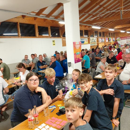 Eine Gruppe von Menschen sitzt in einem Restaurant an Tischen, einige mit Brille und in blauen Uniformen. Sie lächeln und scheinen eine Mahlzeit zu genießen. Dahinter sitzen weitere Menschen, und ein Plakat mit rosa Rand ist an der Wand angebracht.