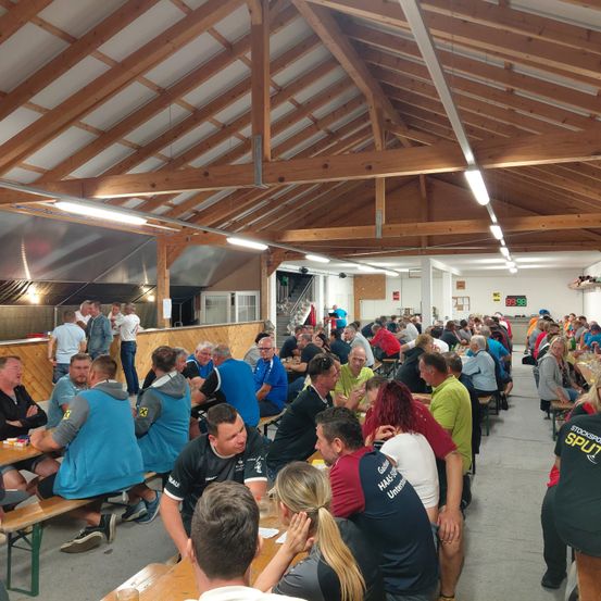 Eine Gruppe von Menschen sitzt an Tischen in einem großen Saal mit Holzbalken und Lampen. Einige unterhalten sich, während andere essen. Die Tische sind mit Tellern und Tassen bedeckt.