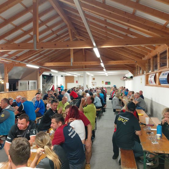 Eine große Gruppe von Menschen sitzt in Reihen in einem Essbereich. Sie führen Gespräche, einige trinken aus Gläsern. Der Raum hat Holzbalken, und auf den Tischen befinden sich verschiedene Gegenstände wie Flaschen und Tassen. An der Wand hängen auch Poster.