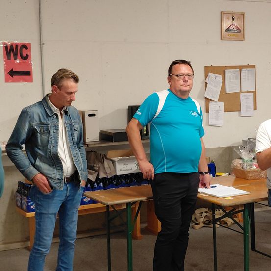 Zwei Männer stehen in einem Raum in der Nähe eines Tisches. Einer trägt eine Jeansjacke und der andere ein blaues Hemd. Papiere und eine Flasche sind auf dem Tisch.