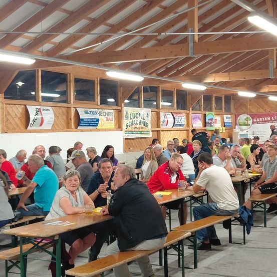 Viele Menschen sitzen auf Bänken in einem Saal, einige unterhalten sich und trinken Bier. Es gibt Banner und Poster an der Wand. Der Saal hat Holzdecken und Fenster.