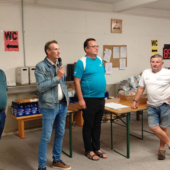 Vier Männer stehen in einem Raum mit einem Tisch vor ihnen. Der Mann in der Mitte hält ein Mikrofon und spricht. Dahinter befindet sich ein Regal mit Lagerboxen und einem gelben Schild, das nach rechts zeigt.