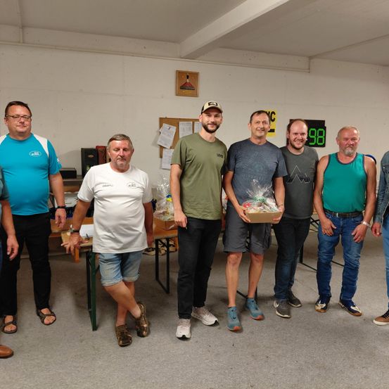 Eine Gruppe von Männern steht in einem Raum, einer hält eine Geschenkbox, lächelt und posiert für ein Foto. Hinter ihnen steht ein Tisch mit einem Lautsprecher und Papieren. Eine digitale Uhr zeigt 98 an.