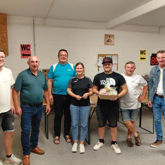 Sieben Personen, bestehend aus Männern und Frauen, versammeln sich in einem Raum mit weißer Wand. Sie scheinen für ein Foto zu posieren. Ein Mann hält einen Korb, und eine Frau steht neben ihm. Hinter ihnen steht ein Holztisch, und zwei Schilder mit der Aufschrift WC sind an der Wand befestigt.