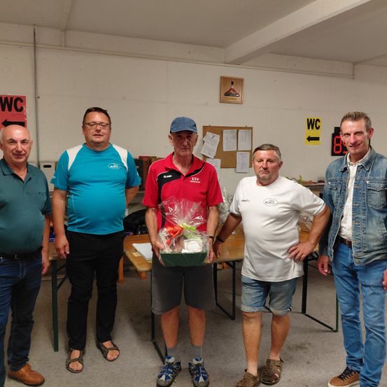 Fünf Männer stehen in einem Raum mit einer weißen Wand und einem WC-Schild. Ein Mann hält eine Geschenkbox, während die anderen lächeln. Hinter ihnen steht ein Tisch mit einer Box darauf.