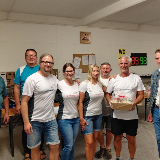 Eine Gruppe von sechs Personen in identischen weißen Shirts und Jeansshorts posiert für ein Foto in einem Raum mit weißen Wänden und einer digitalen Uhr, die 89:98 anzeigt.