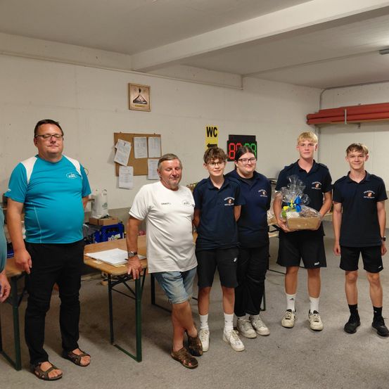 Fünf Personen stehen in einem Raum, möglicherweise einem Club, wobei eine Person einen Geschenkkorb hält. Dahinter ist eine Tafel mit einem digitalen Timer an der Wand montiert.