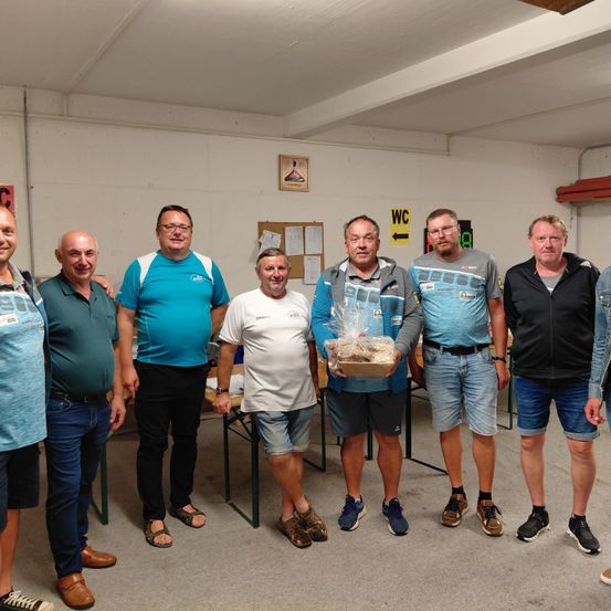 Sieben Männer posieren für ein Foto in einem Raum mit weißen Wänden. Ein Mann hält ein in Plastik eingewickeltes Geschenk.