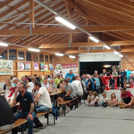 Eine Indoor-Versammlung von Erwachsenen und Kindern, die um Tische herum sitzen, der Raum wird von Deckenleuchten beleuchtet. Ein Coca-Cola-Stand ist im Hintergrund sichtbar.