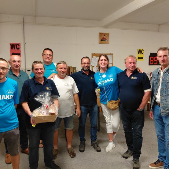 Eine Gruppe von Menschen in blauen Hemden und Sneakers posiert für ein Foto in einem Raum mit WC-Schild und einer Uhr. Eine Frau hält einen Korb mit Essen.
