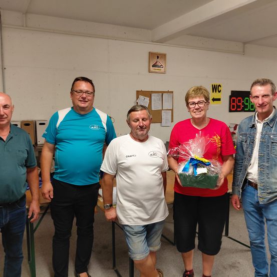 Fünf Erwachsene stehen in einem Raum zusammen, wobei eine Frau einen Geschenkkorb hält. Der Mann in der Mitte trägt ein weißes T-Shirt mit Logo. Hinter ihnen steht ein Tisch und eine Wand mit einem Bilderrahmen und einem Schild.