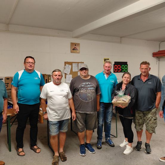 Eine Gruppe von Erwachsenen steht in einem Raum und lächelt für ein Foto. Eine Frau hält einen Geschenkkorb. Hinter ihnen zeigt eine digitale Uhr 89:98. Eine Anschlagtafel und Papiere sind an der Wand angebracht.