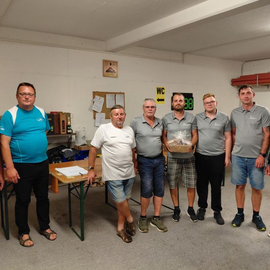 Sechs Männer stehen in einem Raum mit einem Tisch und einer Box in der Mitte. Sie tragen Freizeitkleidung. Eine Tafel und eine Pinnwand sind an der Wand angebracht.