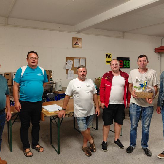 Fünf Männer stehen in einem Raum, drei von ihnen tragen Brillen. Der Mann in der Mitte hält einen Geschenkkorb. Hinter ihnen steht ein Tisch mit einem Notizbuch und einer Schachtel. An der Wand befinden sich ein Anschlagbrett, ein Bilderrahmen und ein Schild mit der Zahl 8:98.