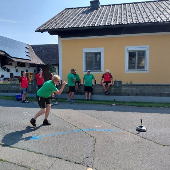 Eine Gruppe von Menschen in grünen und roten Shirts spielt ein Spiel vor einem gelben Haus mit Solarpanelen.