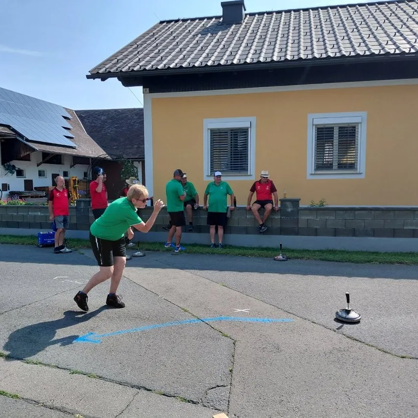 Eine Gruppe von Menschen in grünen und roten Shirts spielt ein Spiel vor einem gelben Haus mit Solarpanelen.