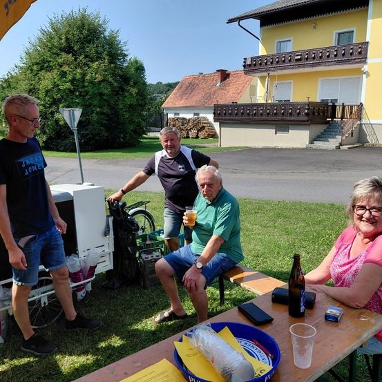 Vier Personen versammeln sich vor einem Haus, zwei sitzen auf einer Bank und zwei stehen. Ein Mann hält ein Getränk, und es gibt einen Tisch mit einer Flasche und anderen Gegenständen. Ein Fahrrad ist in der Nähe geparkt.