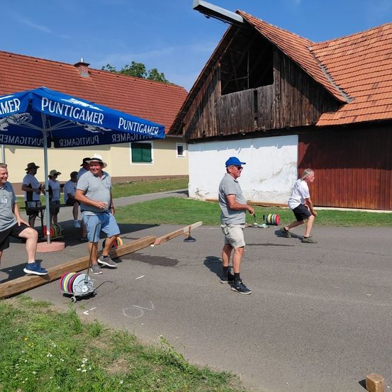 Eine Gruppe von Männern spielt eine Partie Boccia auf einem Betonplatz unter einem blauen Regenschirm.