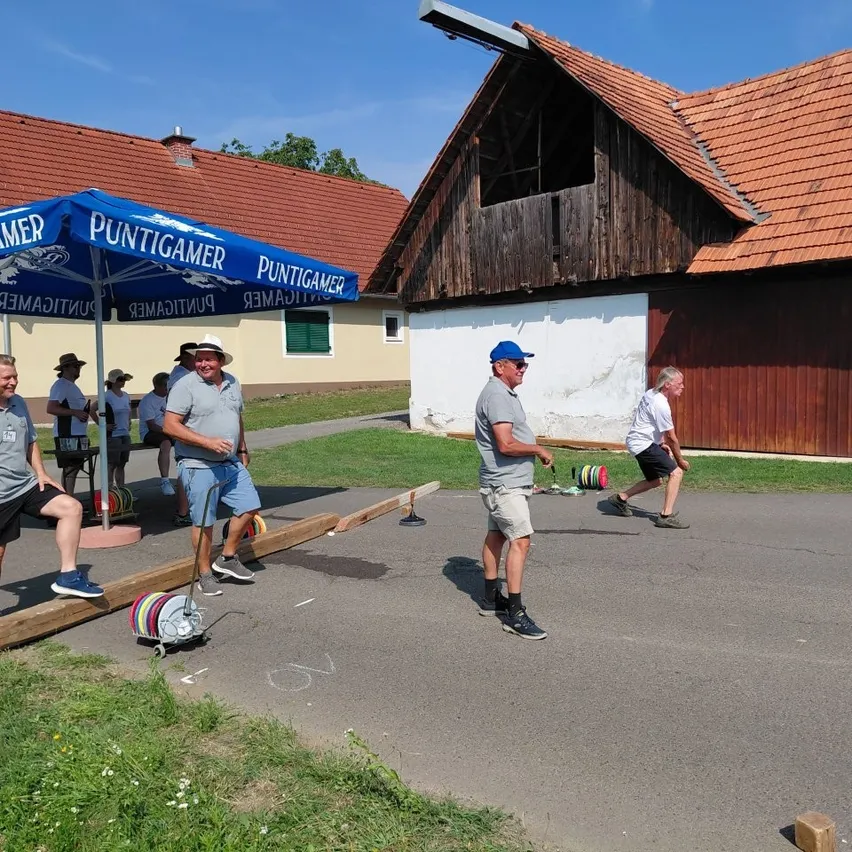 Eine Gruppe von Männern spielt eine Partie Boccia auf einem Betonplatz unter einem blauen Regenschirm.