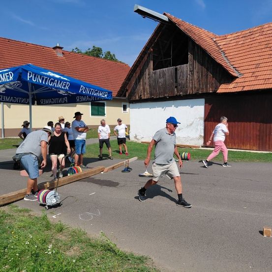 Eine Gruppe von Menschen spielt draußen vor einem Gebäude mit rotem Dach eine Kegelspiel.