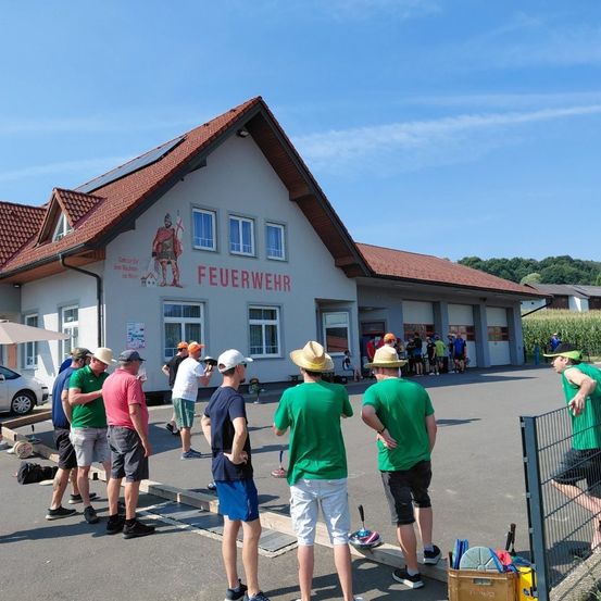 Eine Gruppe von Menschen in grünen Shirts und Hüten steht vor einem Gebäude mit dem Wort 'Feuerwehr' darauf. Sie befinden sich auf einem Parkplatz mit einem geparkten Auto in der Nähe.