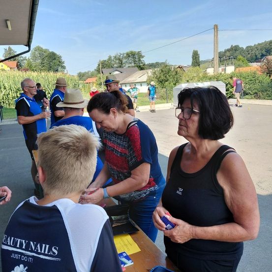 Eine Gruppe von Menschen versammelt sich um einen Tisch, wobei eine Frau in einem rot-blauen Shirt mit einem jungen Jungen in einem blau-weißen Shirt interagiert. In der Nähe schaut eine Frau in einem schwarzen Tanktop zu. Die Szene spielt sich im Freien ab, mit einem Maisfeld und Häusern im Hintergrund.