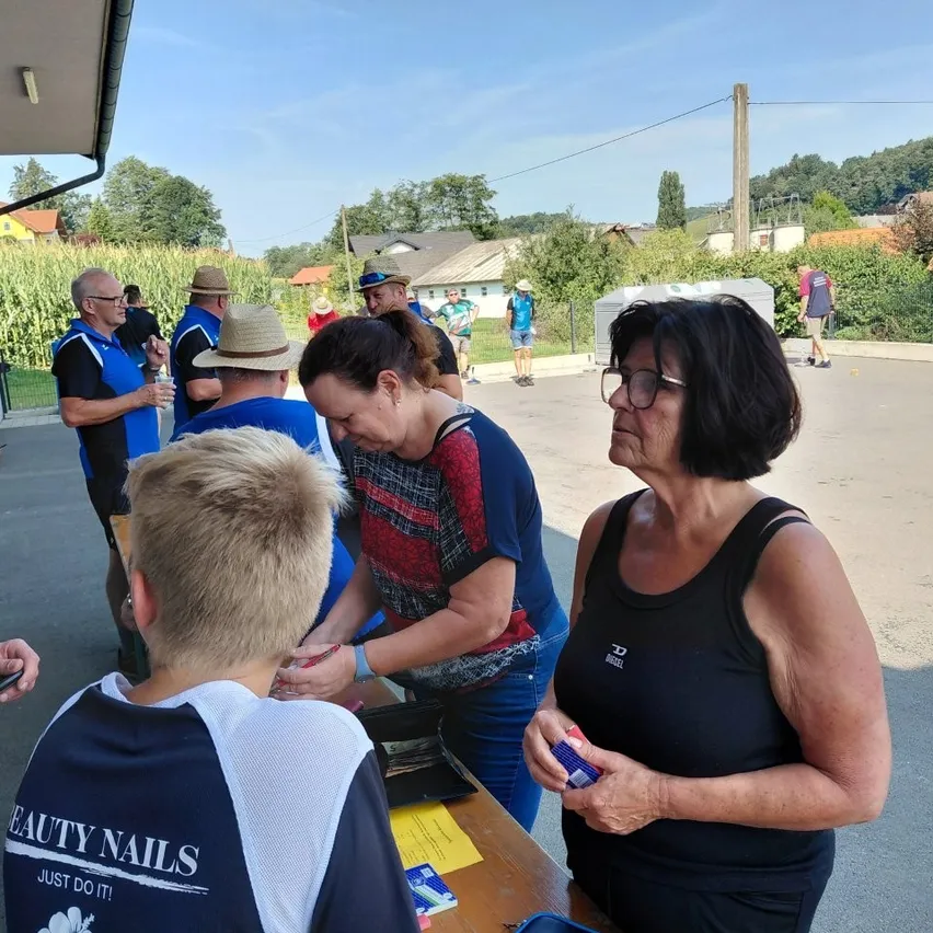 Eine Gruppe von Menschen versammelt sich um einen Tisch, wobei eine Frau in einem rot-blauen Shirt mit einem jungen Jungen in einem blau-weißen Shirt interagiert. In der Nähe schaut eine Frau in einem schwarzen Tanktop zu. Die Szene spielt sich im Freien ab, mit einem Maisfeld und Häusern im Hintergrund.