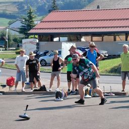 Eine Gruppe von Menschen versammelt sich auf einer Straße und beobachtet, wie ein Mann einen Ball auf ein Ziel wirft. Der Mann trägt ein farbenfrohes Shirt und eine Sonnenbrille. Hinter ihm stehen mehrere Menschen und beobachten, einige tragen Hüte. Im Hintergrund ist ein Gebäude mit einem roten Dach zu sehen.