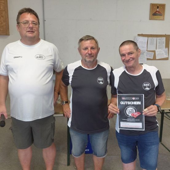 Drei Männer stehen zusammen in einem Raum und lächeln für ein Foto. Der Mann auf der linken Seite hält ein schwarzes Objekt. Der Mann in der Mitte trägt ein schwarz-weißes T-Shirt mit einem Logo. Der Mann auf der rechten Seite hält ein Zertifikat. Hinter ihnen befindet sich ein Bulletin-Board mit Papieren und einem gerahmten Bild.