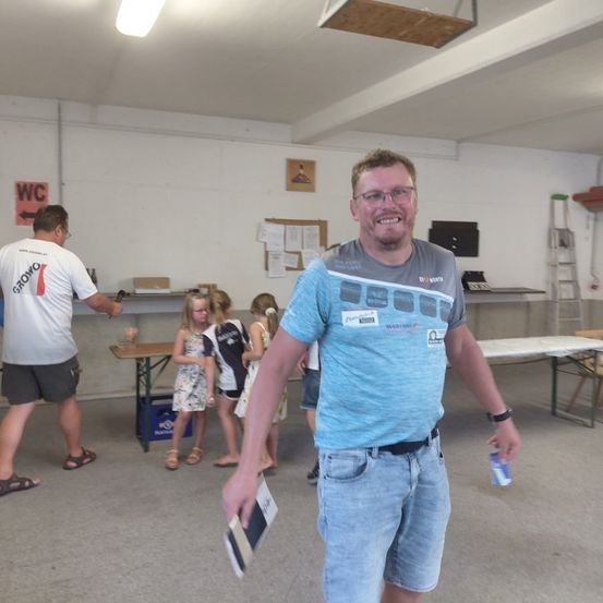 Ein Mann in einem blauen T-Shirt steht in einem Raum mit einer Gruppe von Kindern, lächelt und hält ein Buch. Hinter ihm geht ein Mann in einem weißen T-Shirt. Der Raum hat Tische, Stühle und eine Leiter.