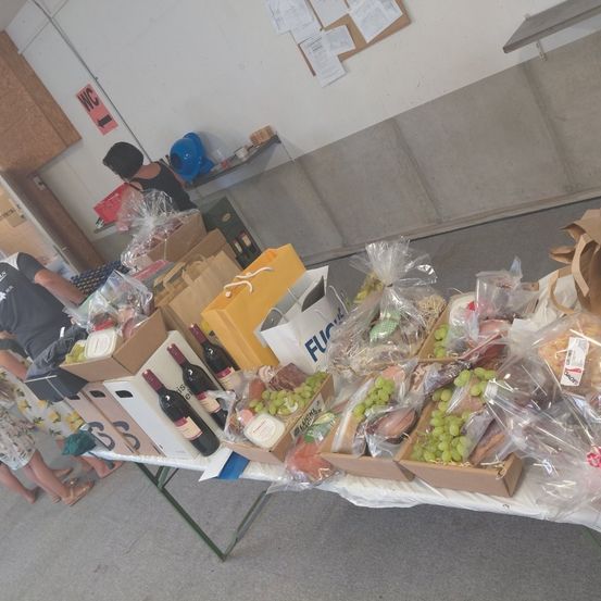 Ein Tisch voller verschiedener Lebensmittel in Kisten und Plastiktüten, darunter Weinflaschen, Trauben und Sandwiches. Zwei Personen stehen hinter dem Tisch, eine trägt ein schwarzes Shirt und die andere ein weißes Kleid.