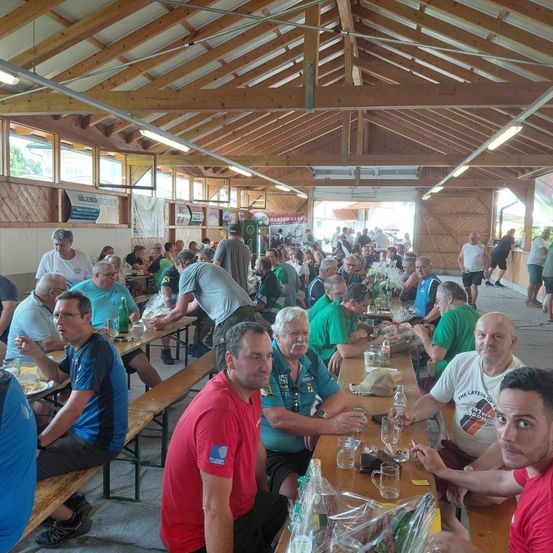 Eine Gruppe von Menschen sitzt an einem langen Tisch in einem Pavillon, einige tragen grüne Shirts, andere rot. Flaschen, Gläser und Essen sind auf dem Tisch. Der Pavillon hat Holzbalken und Glasfenster.