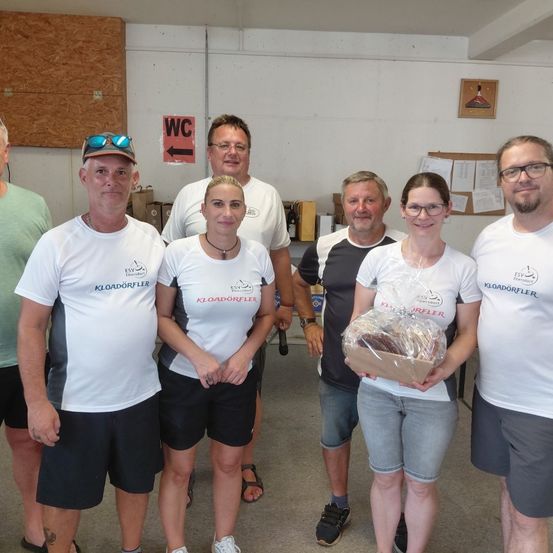 Eine Gruppe von Menschen in passenden weißen T-Shirts posiert für ein Foto. Eine Frau hält ein verpacktes Geschenk. Hinter ihnen hat eine Wand ein Schild, das auf die Toilette weist.