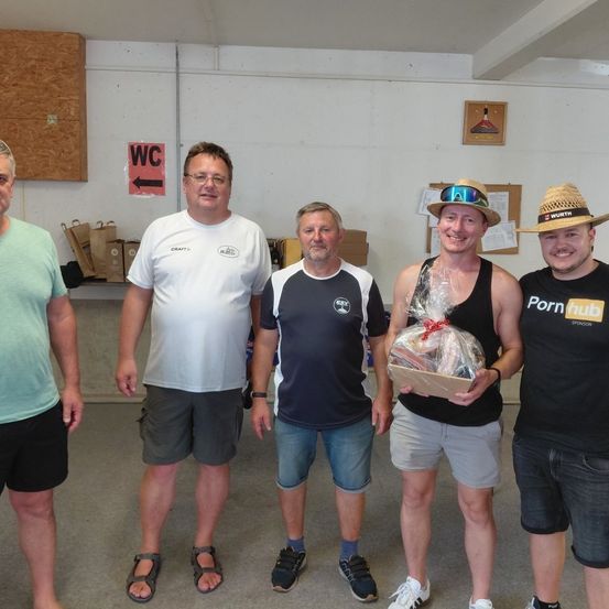 Fünf Männer stehen lächelnd in einem Raum. Ein Mann hält eine Geschenkbox in Plastikfolie. Der Mann ganz links trägt ein grünes Shirt und der Mann ganz rechts trägt einen Hut.