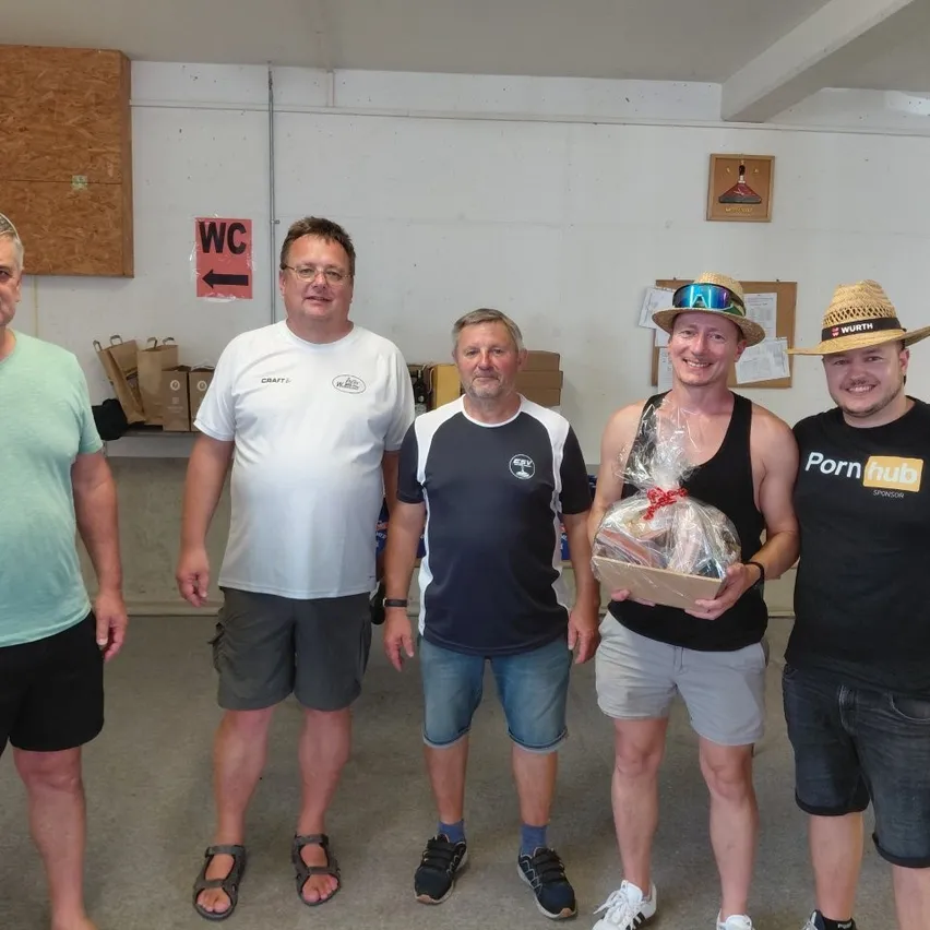 Fünf Männer stehen lächelnd in einem Raum. Ein Mann hält eine Geschenkbox in Plastikfolie. Der Mann ganz links trägt ein grünes Shirt und der Mann ganz rechts trägt einen Hut.