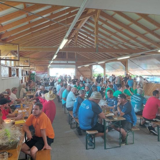 Eine große Gruppe von Menschen sitzt an Tischen in einer großen Holzstruktur. Sie tragen verschiedene farbige Hemden und Hüte. Einige trinken aus Gläsern, andere essen. Der Bereich ist gut beleuchtet mit Holzbalken und Lampen an der Decke.