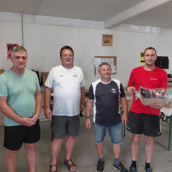 Vier Männer stehen in einem Raum und lächeln für ein Foto. Ein Mann hält einen Korb mit Flaschen. Der Raum hat weiße Wände und einen Tisch mit Gegenständen darauf.