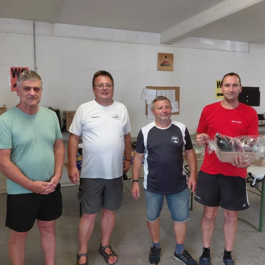 Vier Männer stehen in einem Raum und lächeln für ein Foto. Ein Mann hält einen Korb mit Flaschen. Der Raum hat weiße Wände und einen Tisch mit Gegenständen darauf.