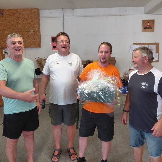 Vier Männer in Freizeitkleidung stehen in einem Raum, einer hält ein verpacktes Geschenk. Der Mann in der Mitte trägt ein orangefarbenes Shirt, die anderen tragen weiße, grüne und schwarze Shirts. Hinter ihnen stehen Lagerboxen und eine Pinnwand.