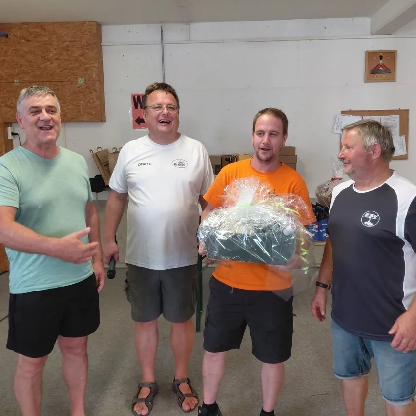 Vier Männer in Freizeitkleidung stehen in einem Raum, einer hält ein verpacktes Geschenk. Der Mann in der Mitte trägt ein orangefarbenes Shirt, die anderen tragen weiße, grüne und schwarze Shirts. Hinter ihnen stehen Lagerboxen und eine Pinnwand.