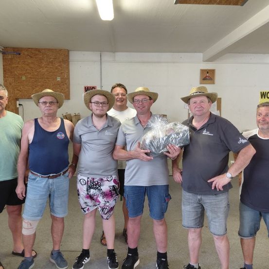 Eine Gruppe von Männern in Strohhüten und lässiger Kleidung steht in einem Raum, einer hält ein verpacktes Geschenk. Sie scheinen ein Ereignis zu feiern.