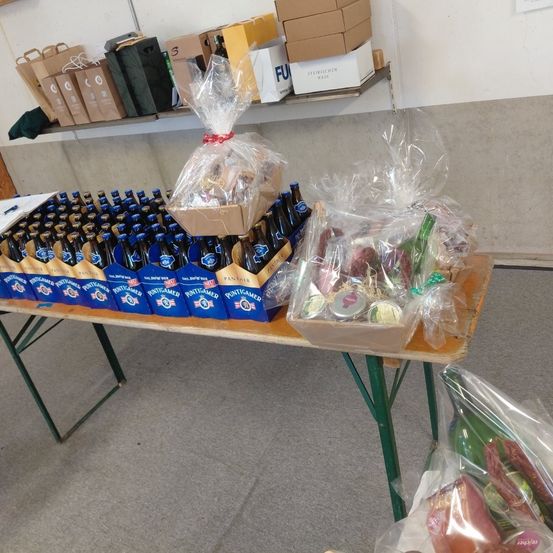 Ein Tisch zeigt verschiedene Geschenkkörbe und Bierflaschen, einige in Plastik verpackt und andere in Kisten. Hinter dem Tisch befinden sich Regale mit Lagerkisten und Taschen.
