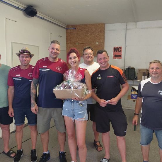 Eine Gruppe von Menschen in einem Raum, lächelnd und für ein Foto posierend. Eine Frau in der Mitte hält eine Geschenkbox. Hinter ihnen weist ein Schild auf die Toilette hin.