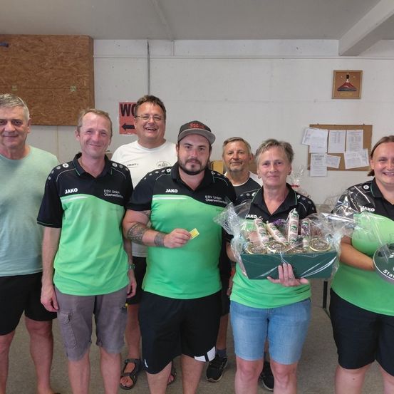 Eine Gruppe von Menschen in grünen und schwarzen Uniformen steht zusammen und lächelt für ein Foto. Sie halten eine Box mit Gegenständen. Der Mann in der Mitte trägt einen Hut. Hinter ihnen befindet sich eine weiße Wand mit einer Pinnwand.