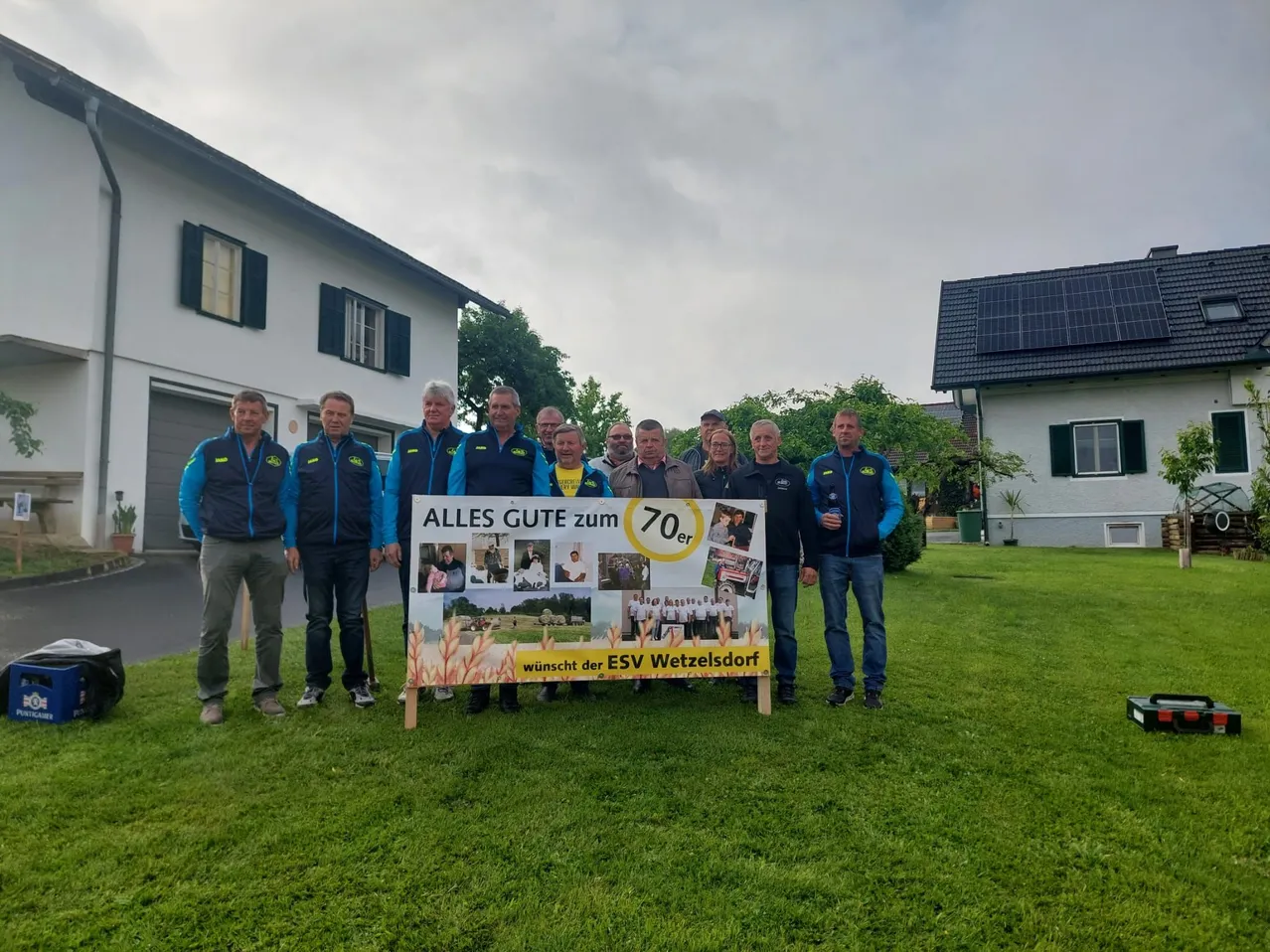 Eine Gruppe von Männern posiert vor einem großen Poster auf einem Rasen. Das Poster hat Bilder von Menschen und den Text 'Alles Gute zum 70er'.