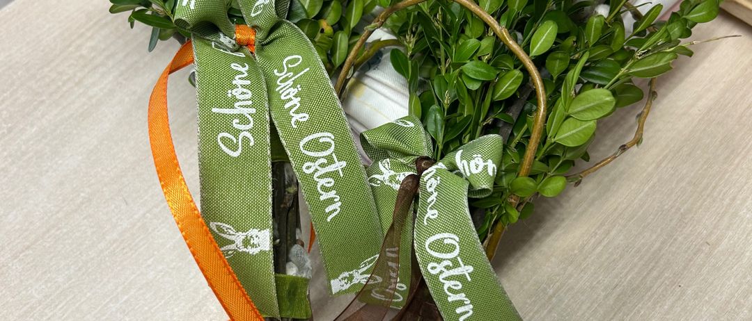 Zwei Ostersträuße mit grünen Bändern, einer mit der Aufschrift 'Schone Ostern' und der andere mit 'Frohe Ostern', stehen auf einem Tisch mit einem weißen Teller und Messer.